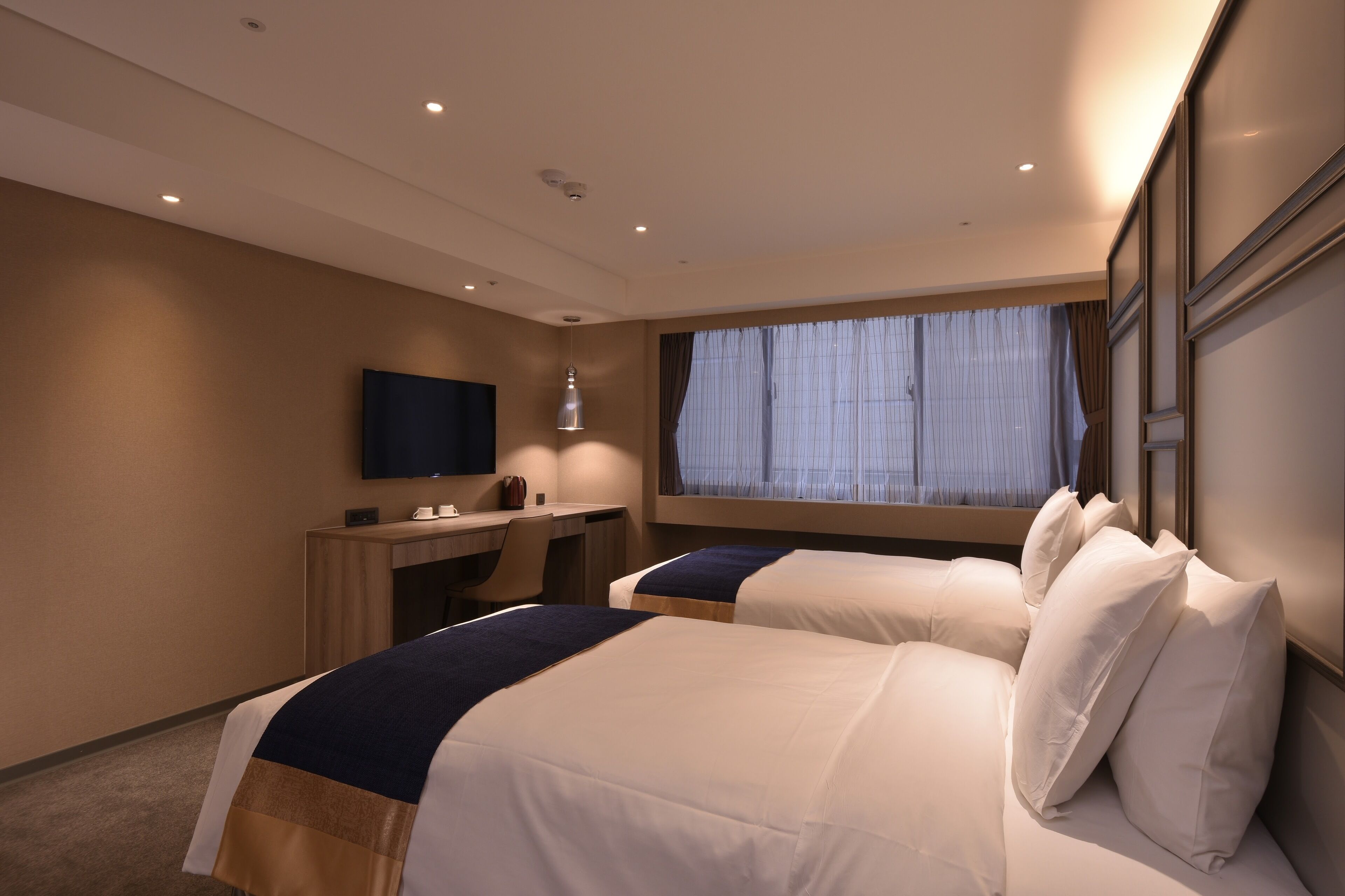 A hotel room with two beds, a desk, a TV, and a window with curtains.