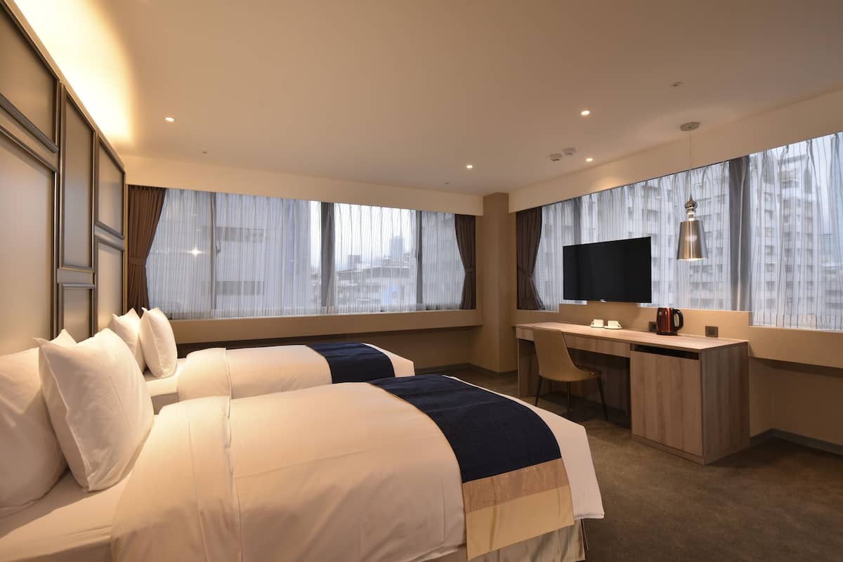 Habitación de hotel con dos camas, un escritorio, una silla, un televisor y ventanas amplias con cortinas translúcidas.