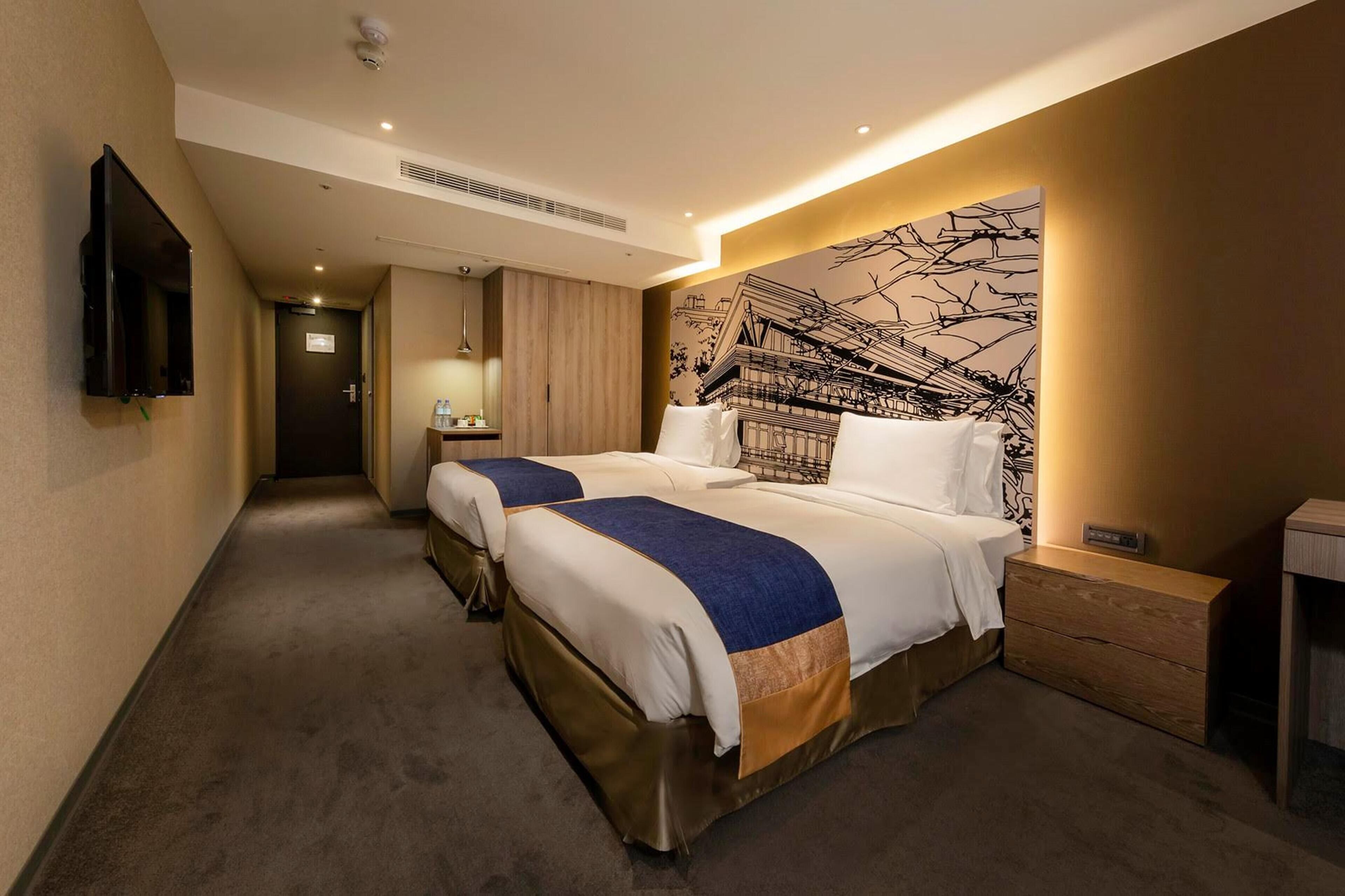 Une chambre d’hôtel avec deux lits, un téléviseur à écran plat fixé au mur, une table de chevet en bois et une grande fresque abstraite sur le mur.
