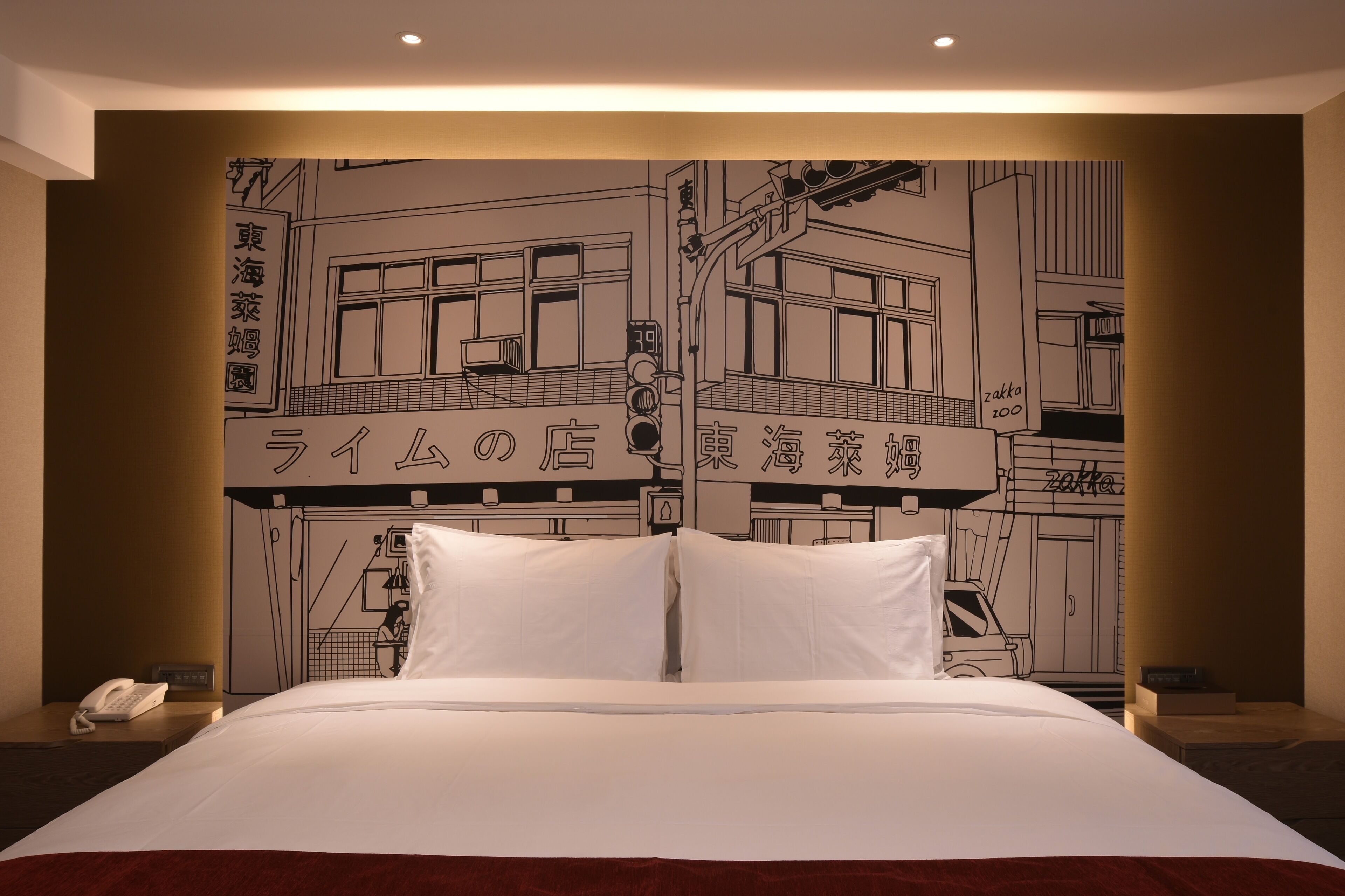 A hotel room with a bed, white pillows, a nightstand with a phone, and a wall with a black and white illustration of a building.