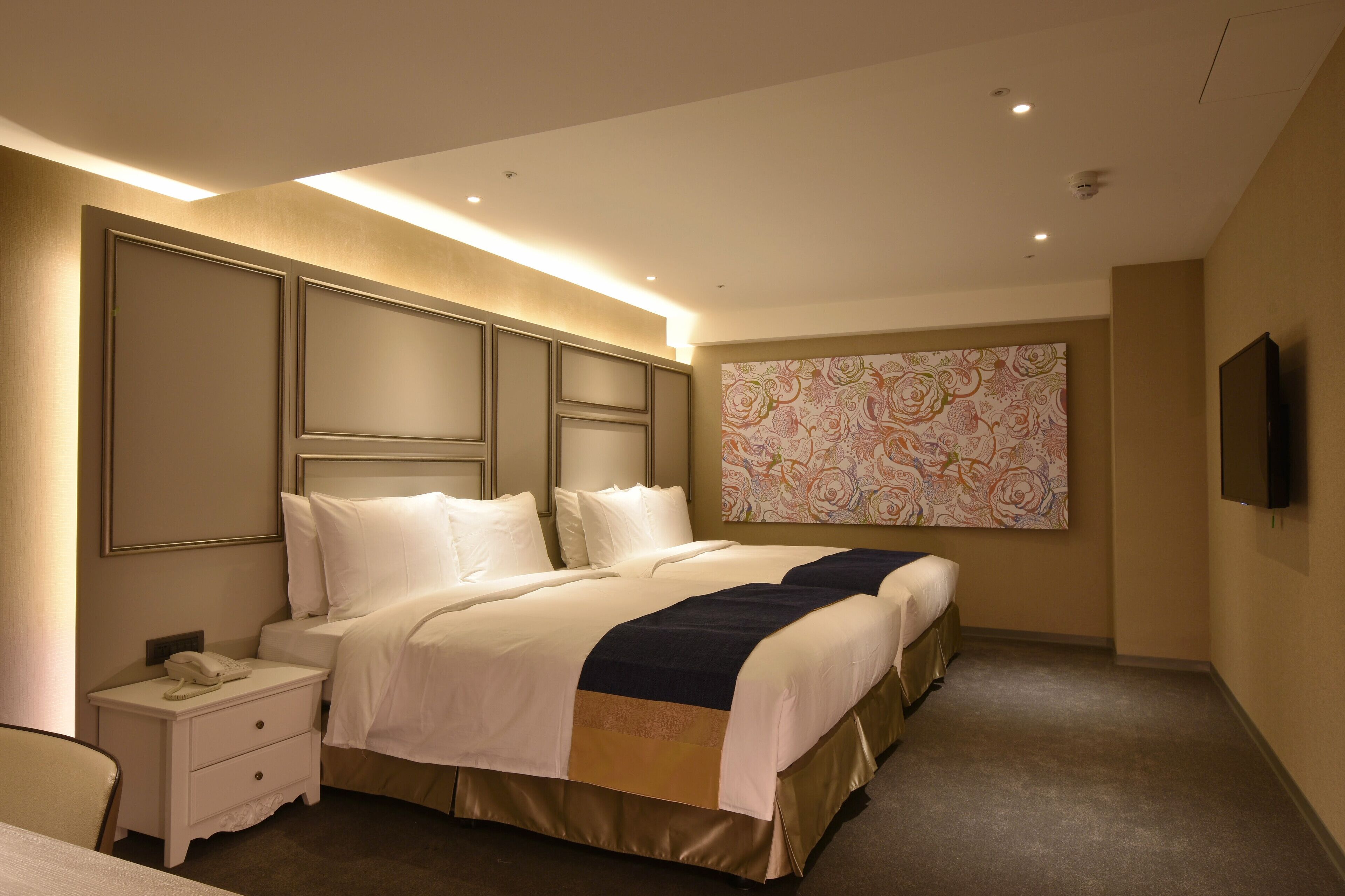 A hotel room with a large bed, a bedside table with a phone, a wall-mounted TV, and a large floral artwork.