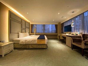 Ein Hotelzimmer mit einem großen Bett, einem Schreibtisch, einem Stuhl, einem Fernseher und einem Fenster mit Vorhängen.