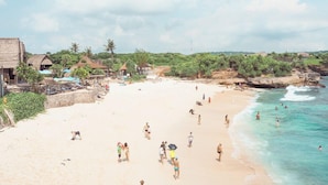 A beach with people swimming and walking on the sand, surrounded by thatched-roof huts and greenery.