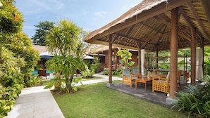 A covered outdoor seating area with wooden furniture, surrounded by lush greenery and a clear sky.