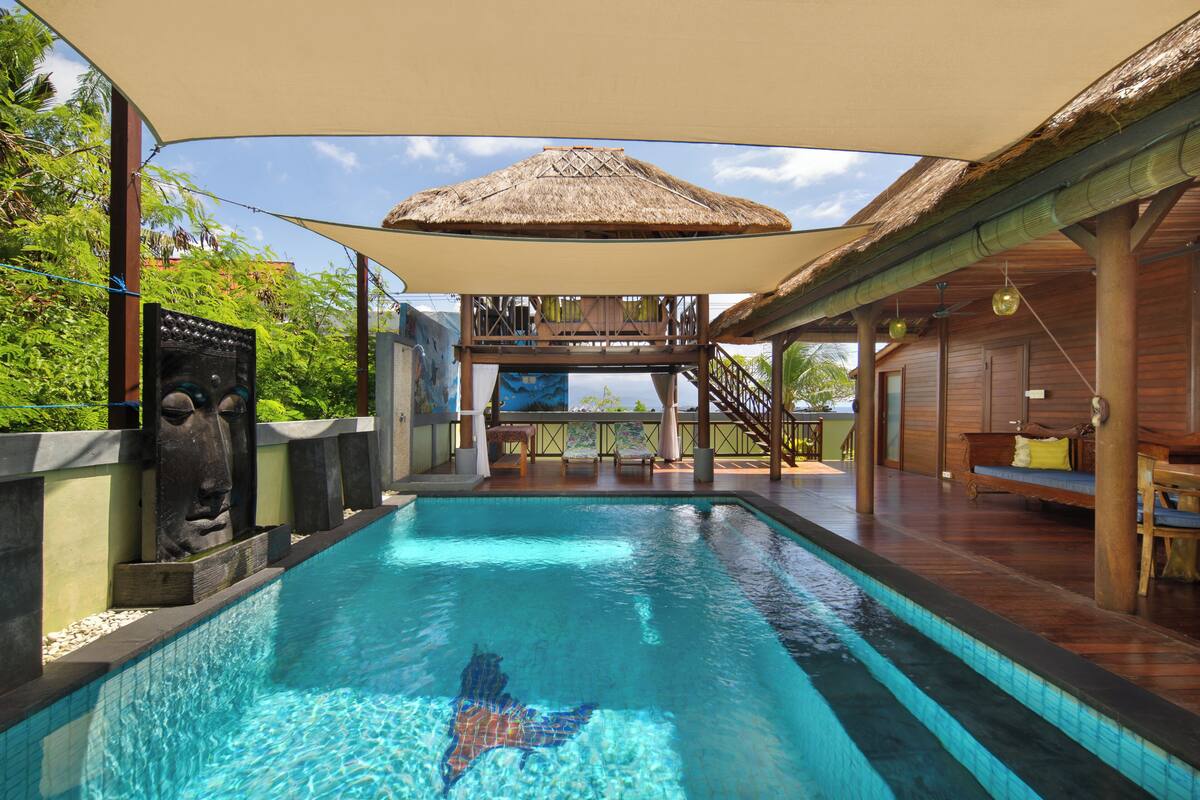 A swimming pool with a thatched-roof structure, wooden deck, and a view of a building with balconies.