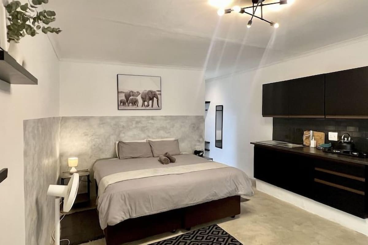 A modern bedroom with a bed, bedside table, and a small desk. There is a kitchen area with cabinets and a countertop.