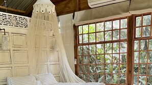 A wooden bed with a canopy, a wicker chair, and a large window with a view of greenery. - LoerieRoep (Mbombela)