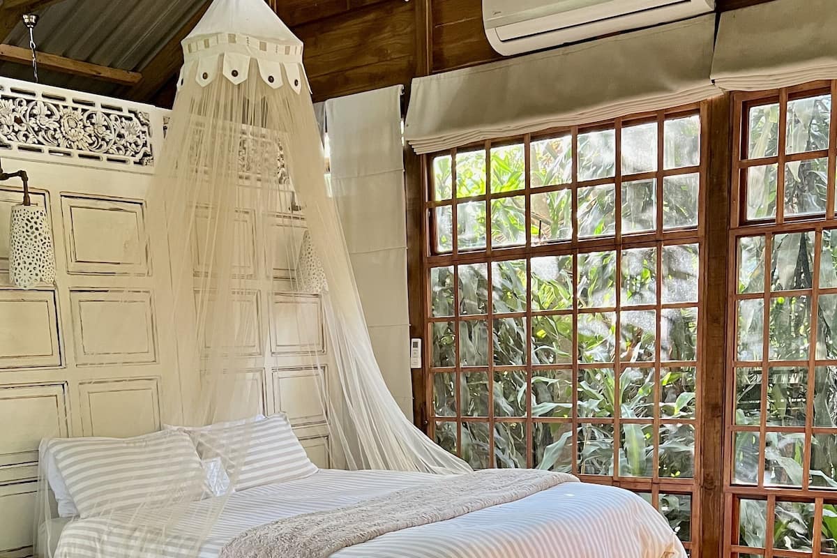 A wooden bed with a canopy, a wicker chair, and a large window with a view of greenery.