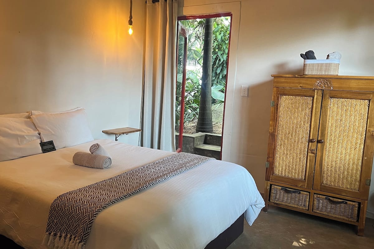 A neatly made bed with a rolled towel, a small bedside table, and a wooden wardrobe with woven doors.