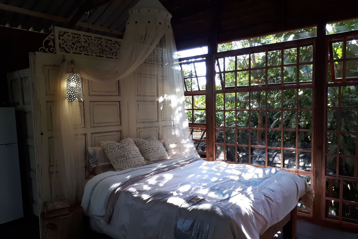 A bedroom with a bed, a nightstand, a decorative wall, and a window with a view of greenery.