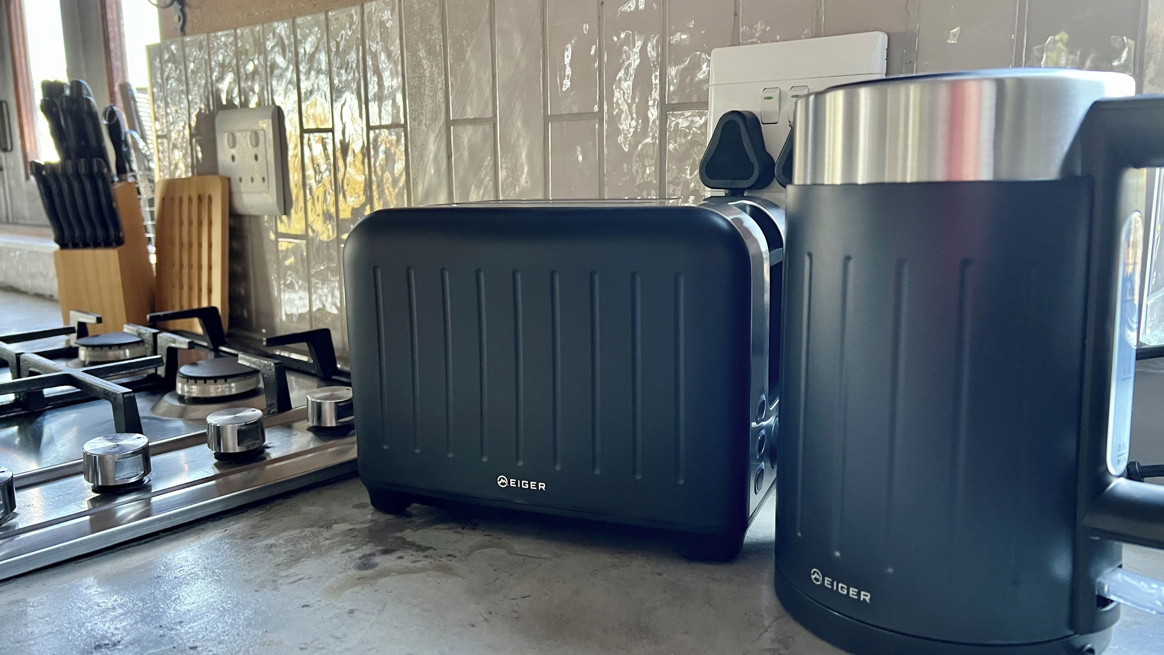 A kitchen with a gas stove, a black Eiger electric kettle, and a black Eiger electric kettle.