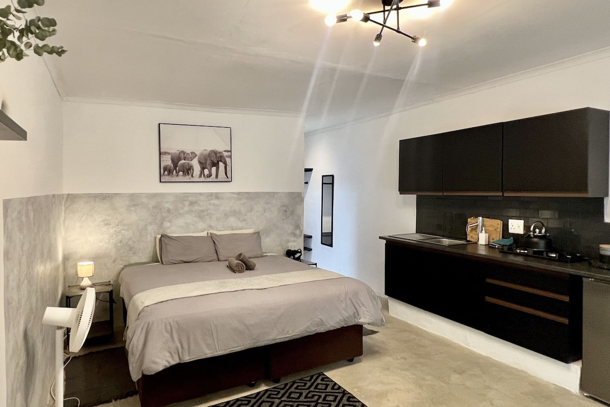 A modern bedroom with a large bed, bedside table, and a nightstand. There is a kitchen area with dark cabinets and a countertop.