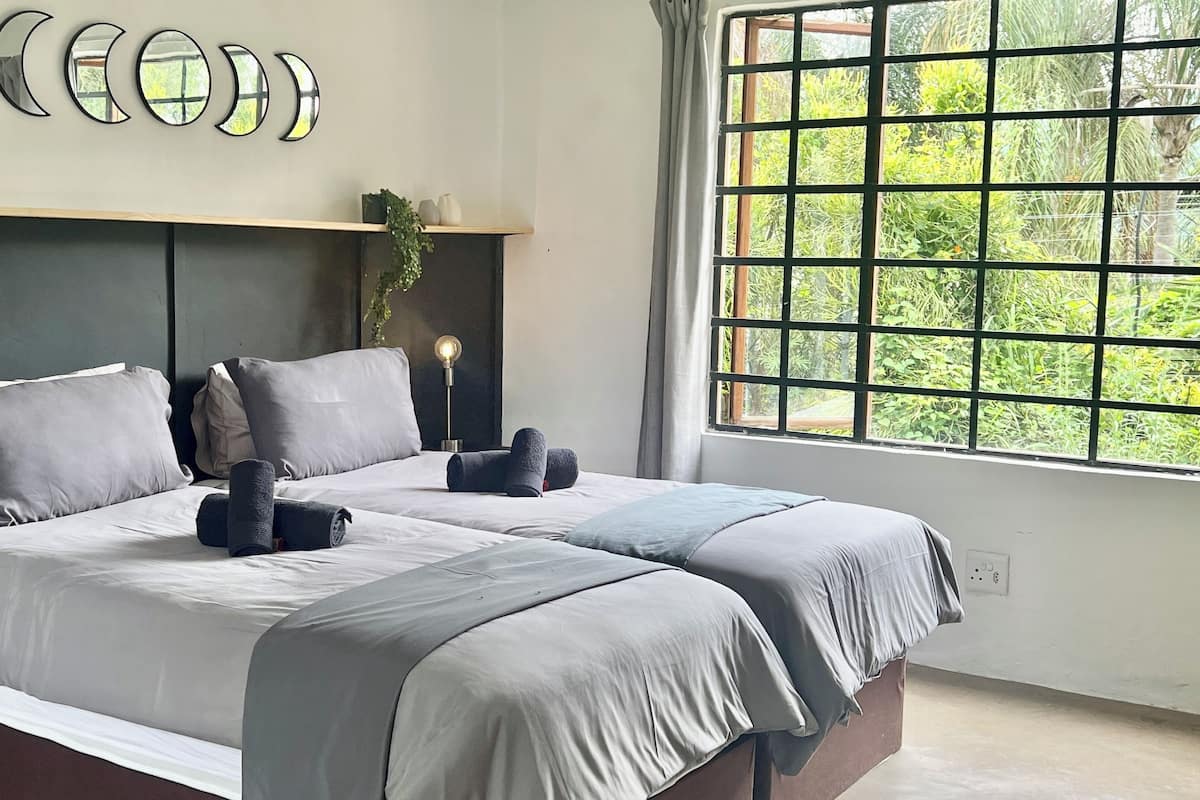 A double bed with grey linens, a bedside lamp, and a view of greenery through a window with a grid pattern.