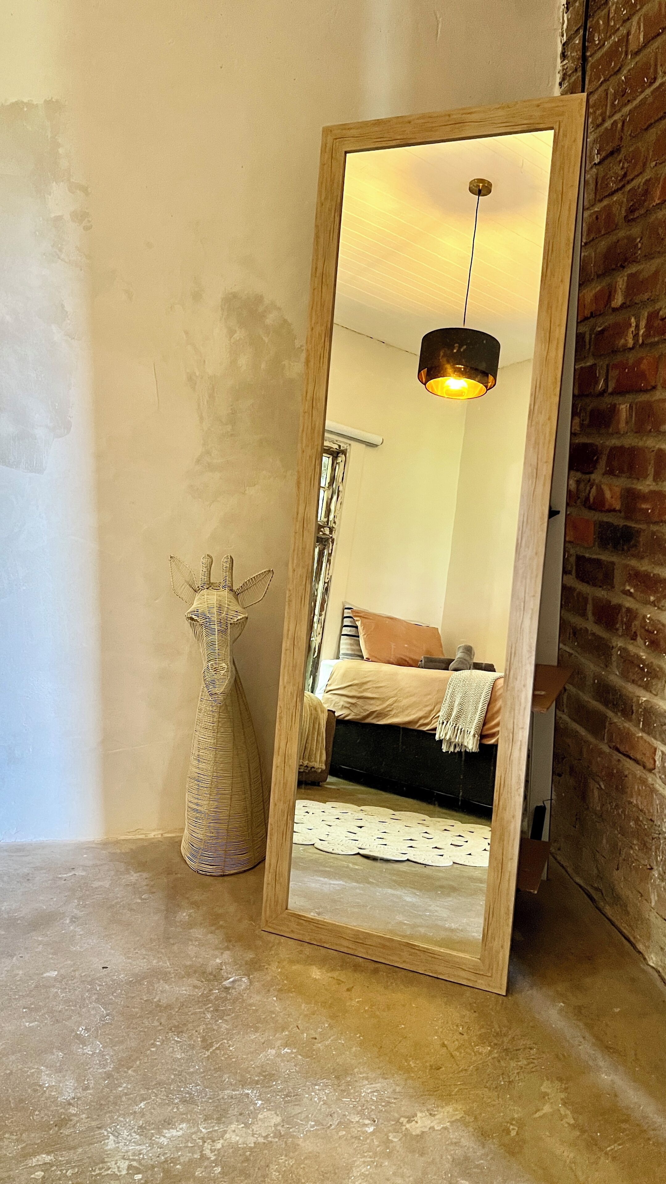 A mirror reflecting a room with a bed, a hanging light fixture, and a wicker vase.