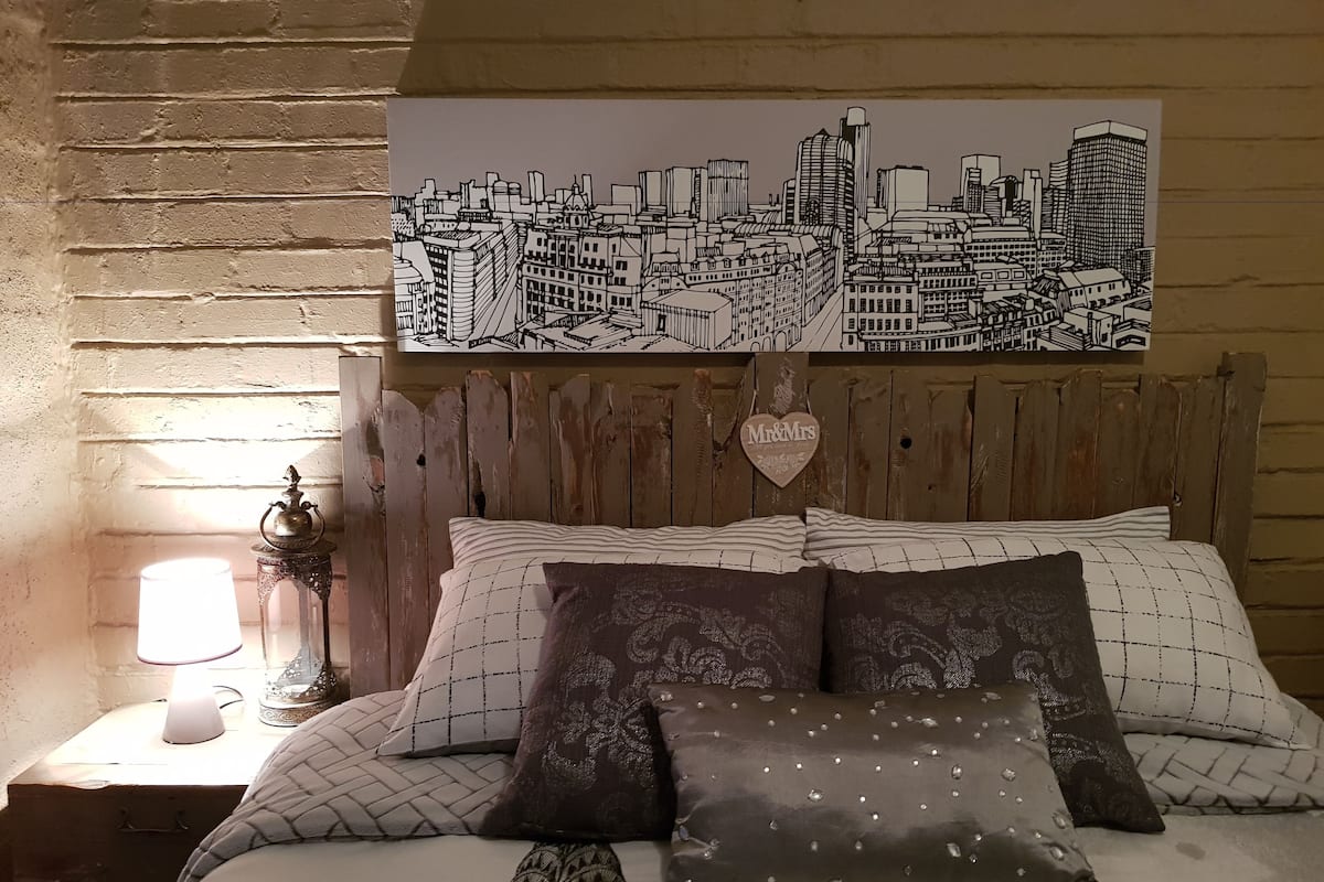 A bedroom with a cityscape artwork, a wooden headboard, patterned bedding, and a bedside table with a lamp.