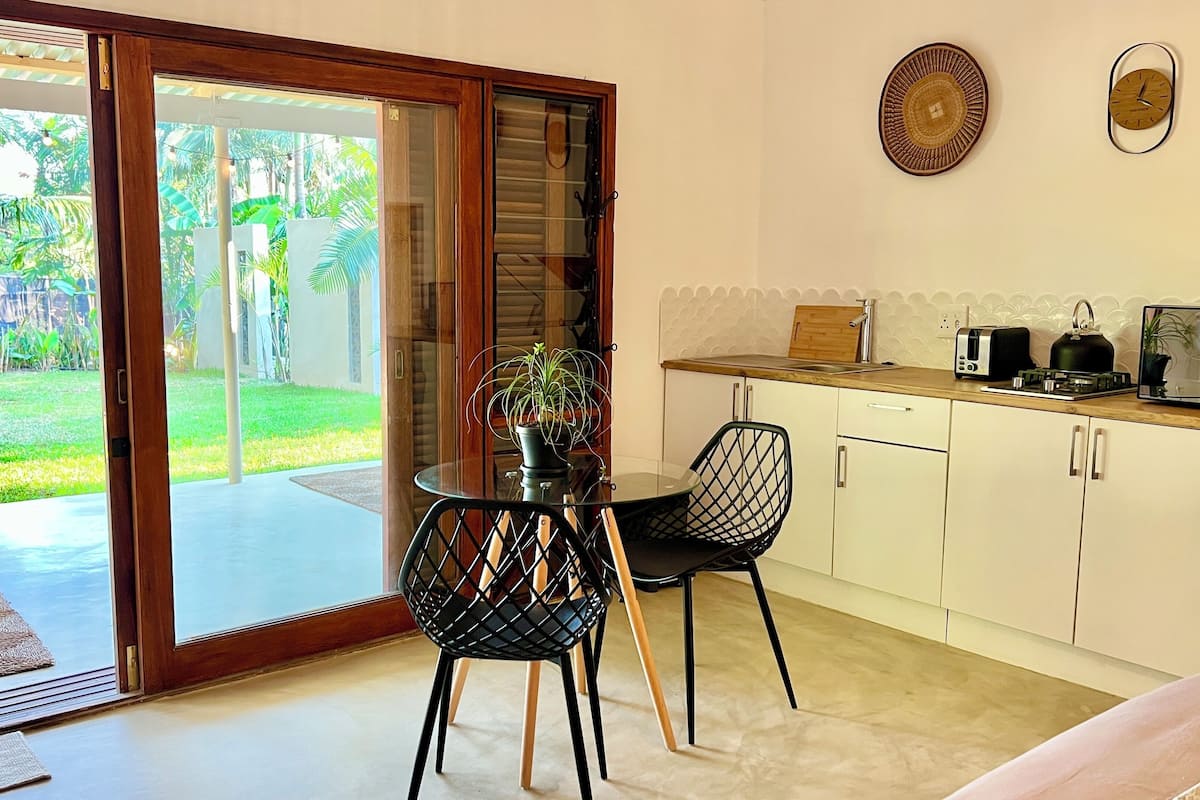 A modern kitchen with a dining area, featuring a glass door leading to a garden view, a round table, and a black chair.
