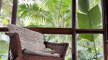 A wicker chair with a crocheted throw, positioned in front of large windows with a view of lush green foliage.
