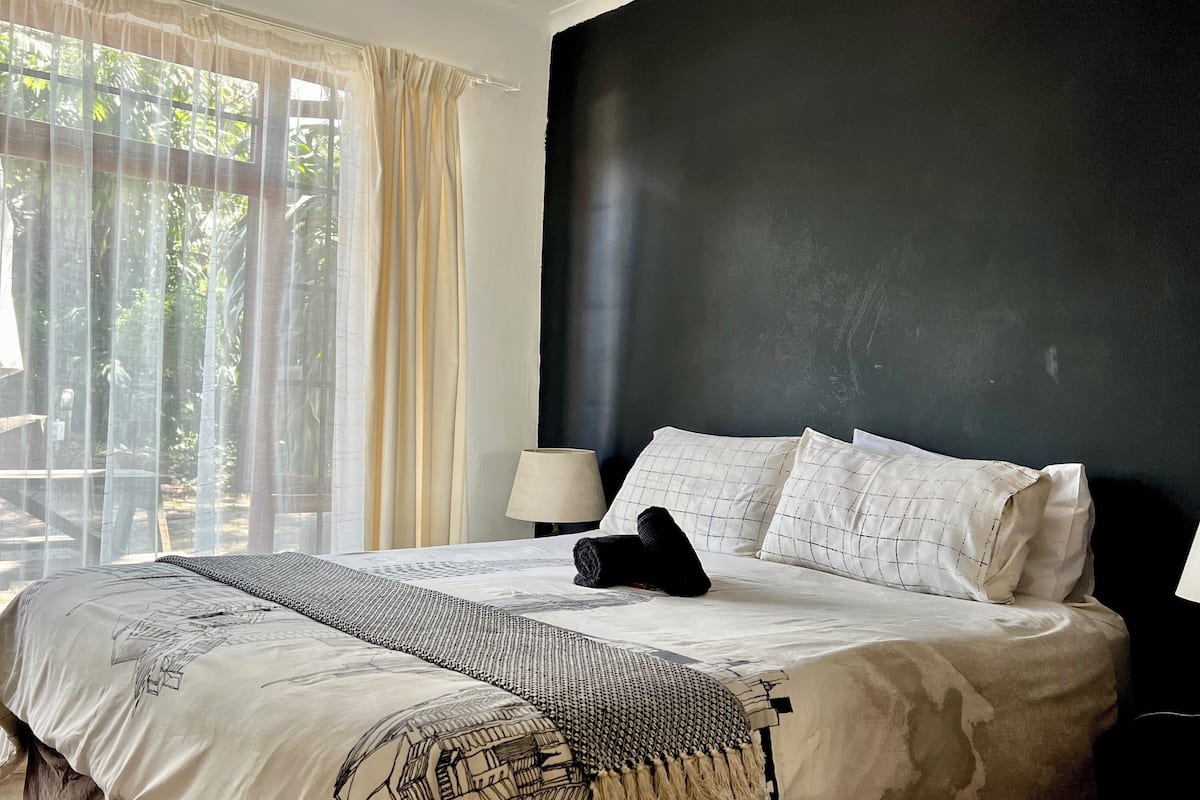 A bedroom with a bed, two bedside lamps, a window with curtains, and a view of greenery outside.