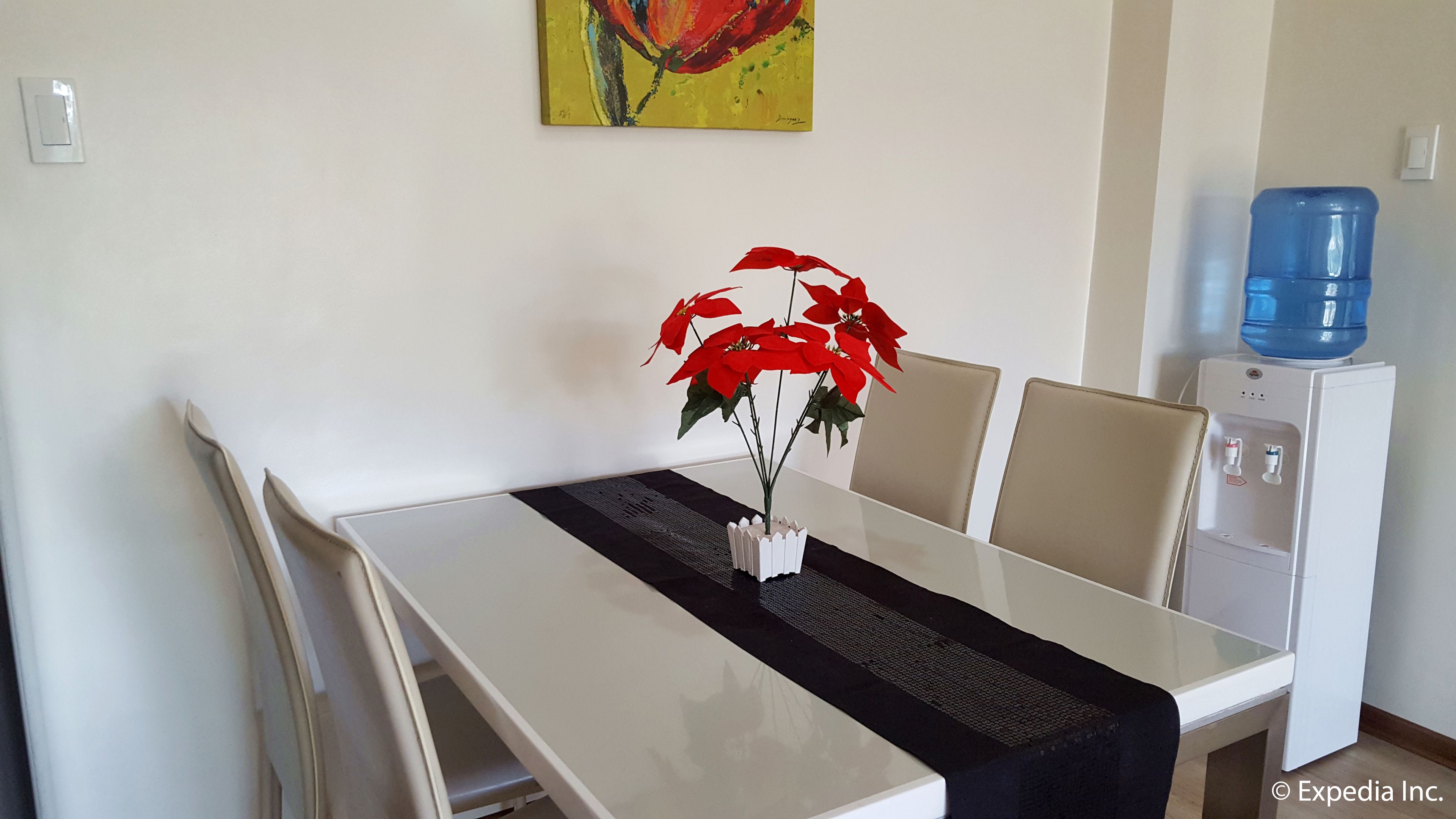 Een eetruimte met een witte tafel, stoelen en een vaas met rode bloemen. Er is een waterdispenser en een schilderij aan de muur.