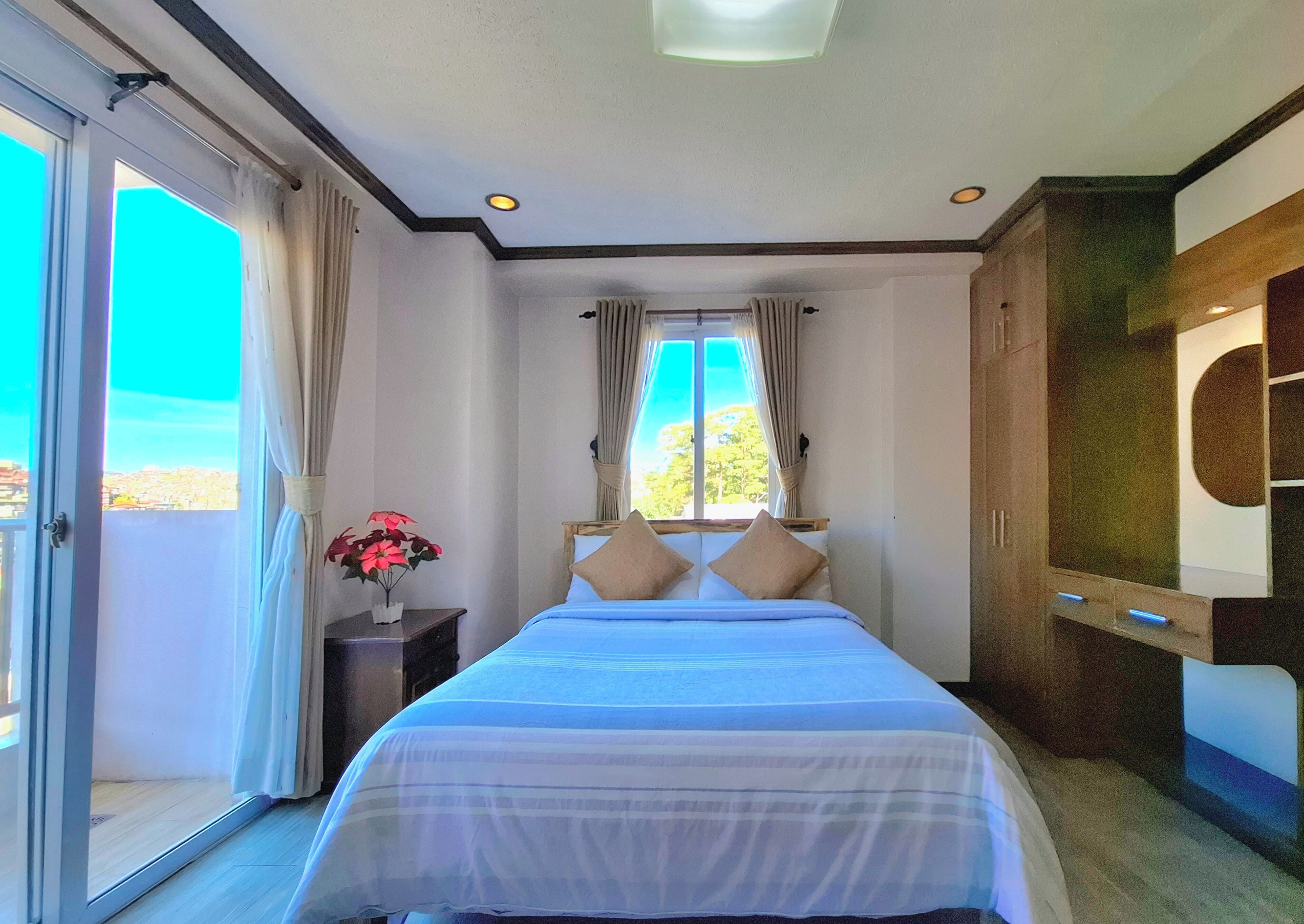 A bedroom with a bed, a nightstand, a potted plant, and a view of the outdoors.