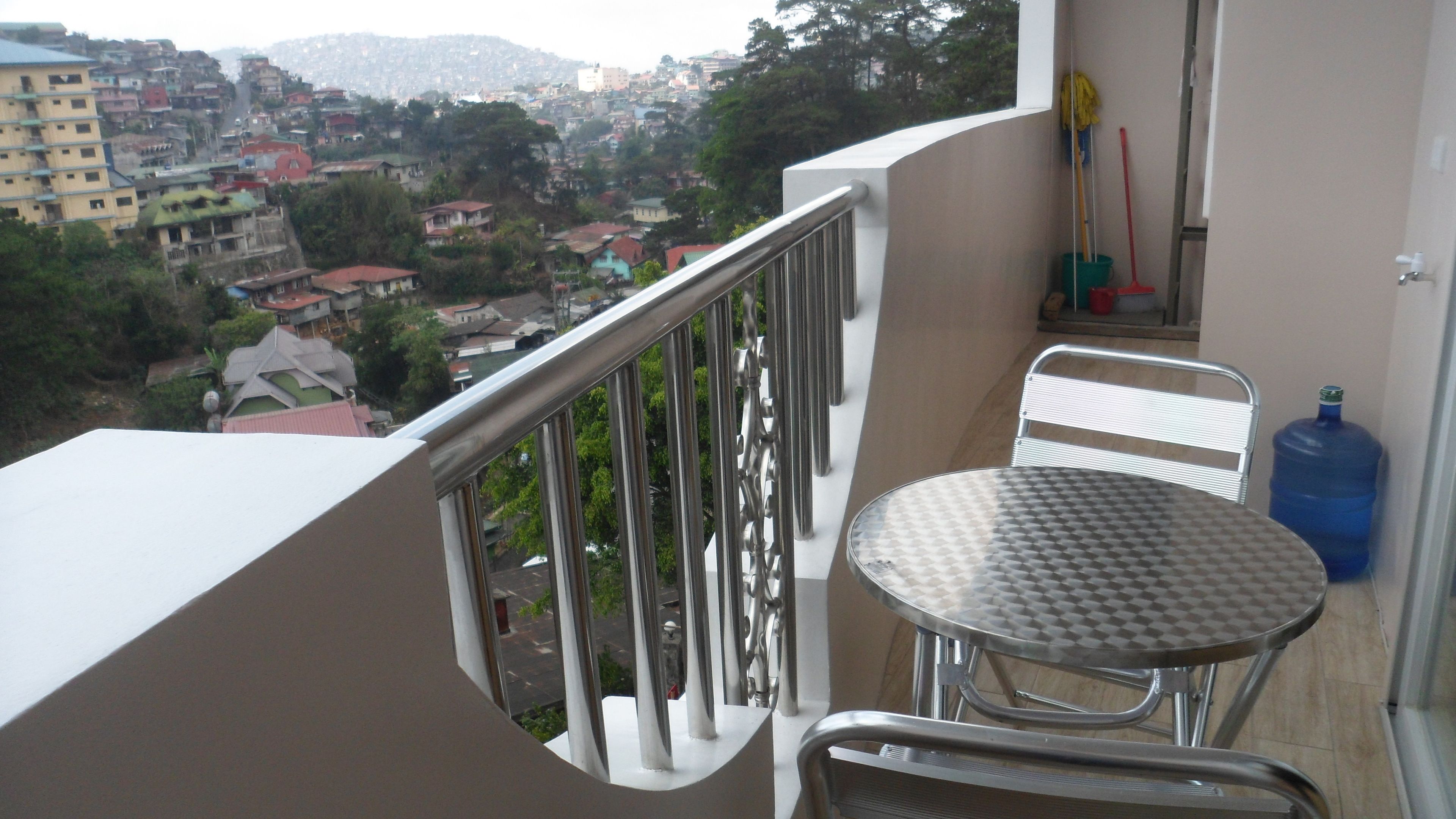 A balcony with a metal railing, a round table, and a chair. There is a large blue water dispenser on the floor.