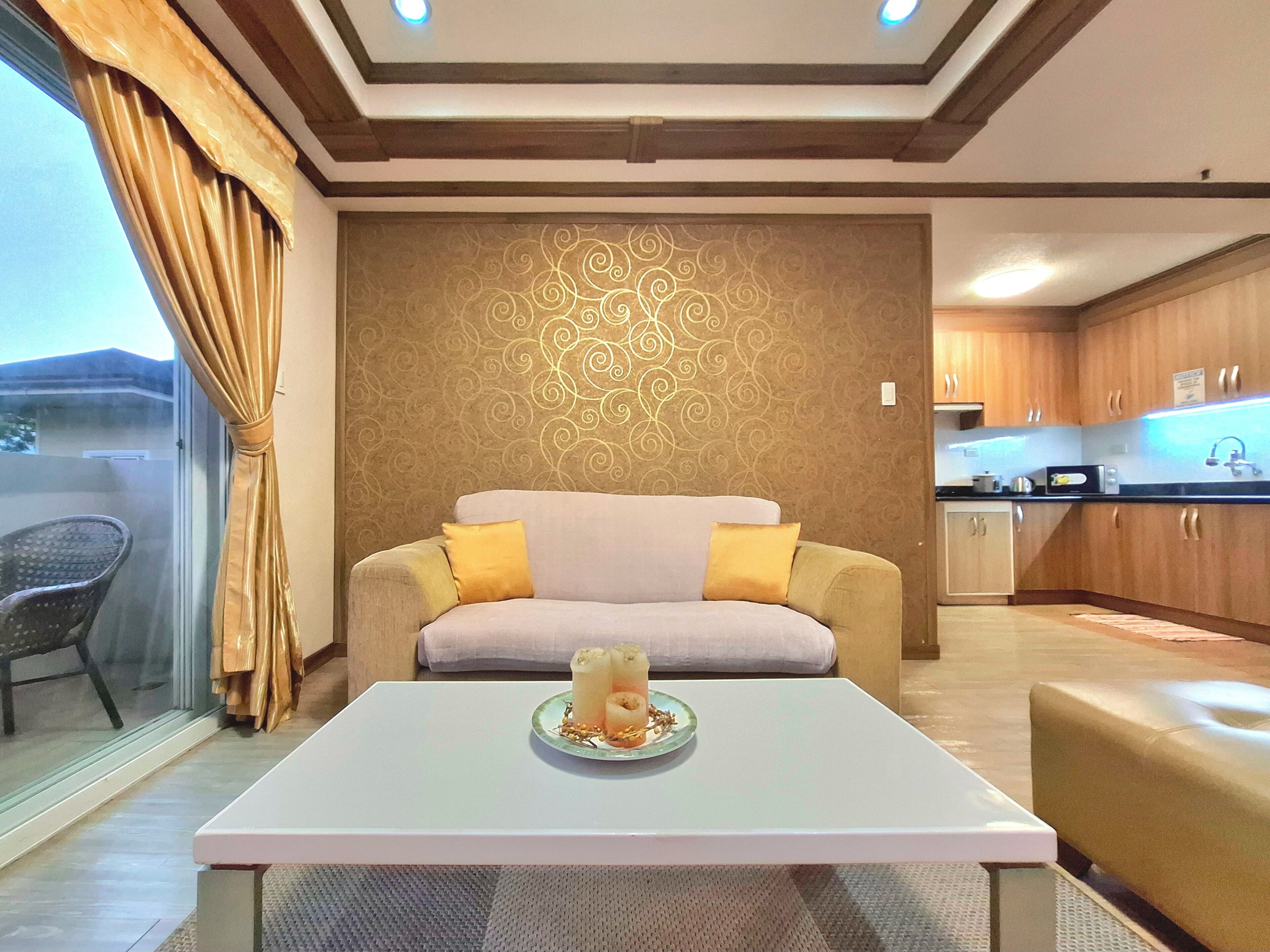 A modern living room with a sofa, a coffee table with a decorative item, a patterned wall, and a kitchen area visible through an open door.
