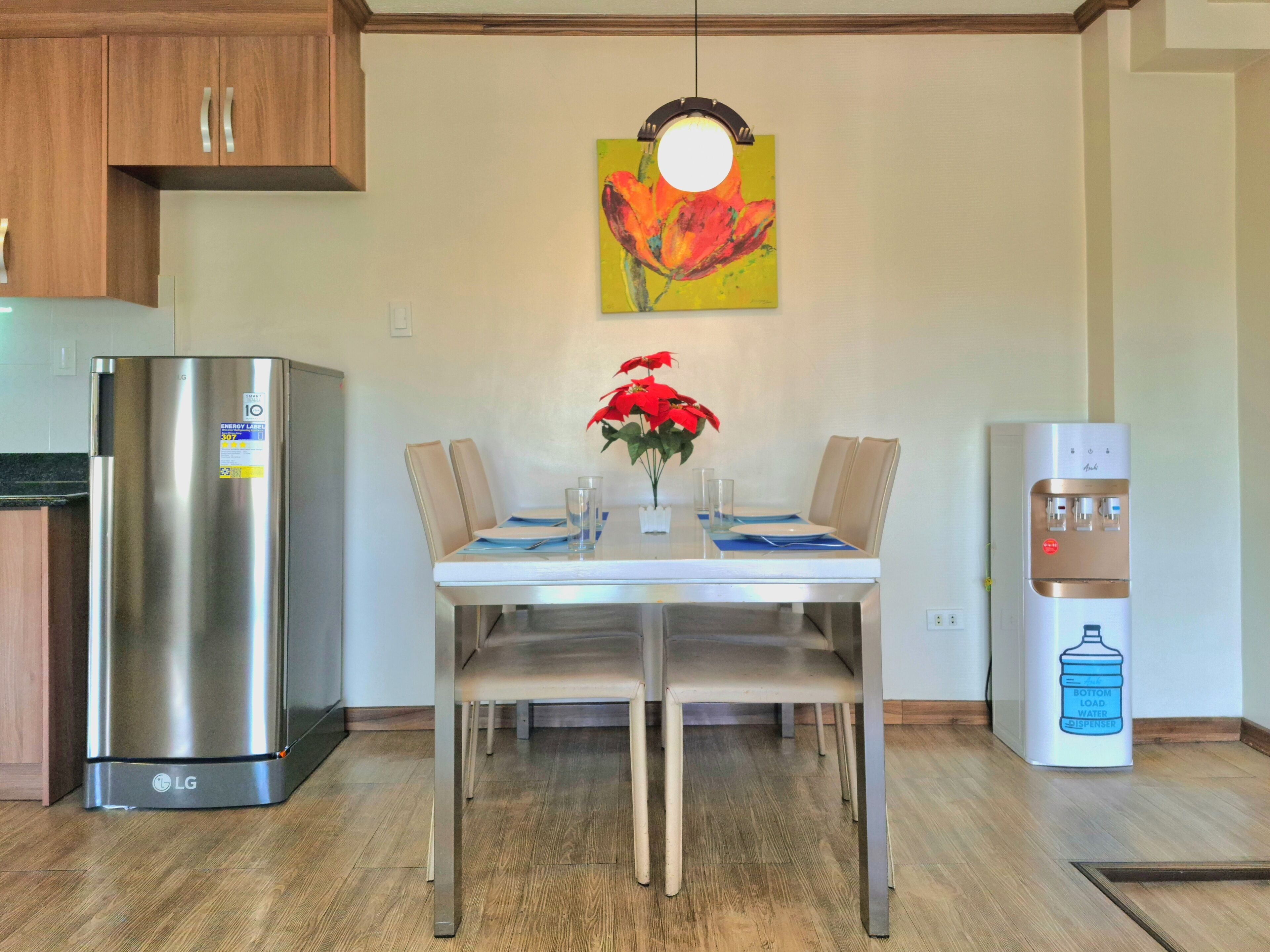 A modern kitchen with a stainless steel refrigerator, a dining table with chairs, a water dispenser, and a colorful painting on the wall.