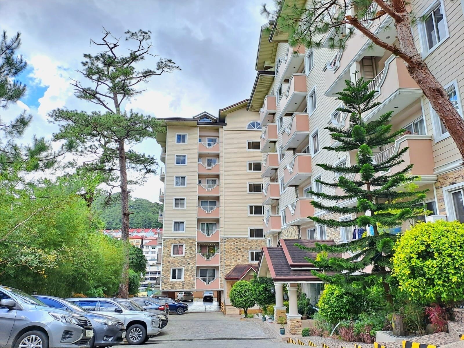 A multi-story residential building with balconies, a parking area, and surrounding greenery.