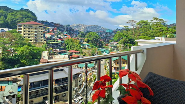 A balcony with a view of a densely built town, a building with a red roof, and a flower arrangement. - Prestige Vacation Apartments - Bonbel Condominium (Baguio)