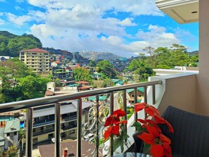 Un balcon donnant sur une ville densément construite, un immeuble avec un toit rouge et un arrangement floral.