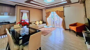 A modern living room with a glass dining table, a sofa, and a red armchair. There is a kitchen area with a sink and cabinets, and a balcony with a view. - Prestige Vacation Apartments - Bonbel Condominium (Baguio)