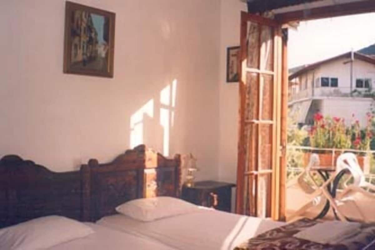 A bedroom with a wooden bed, a small bedside table, a window with a view of a building and flowers outside.