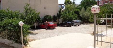 A parking area with a red car and several other vehicles parked. There is a sign indicating the name of the hotel.