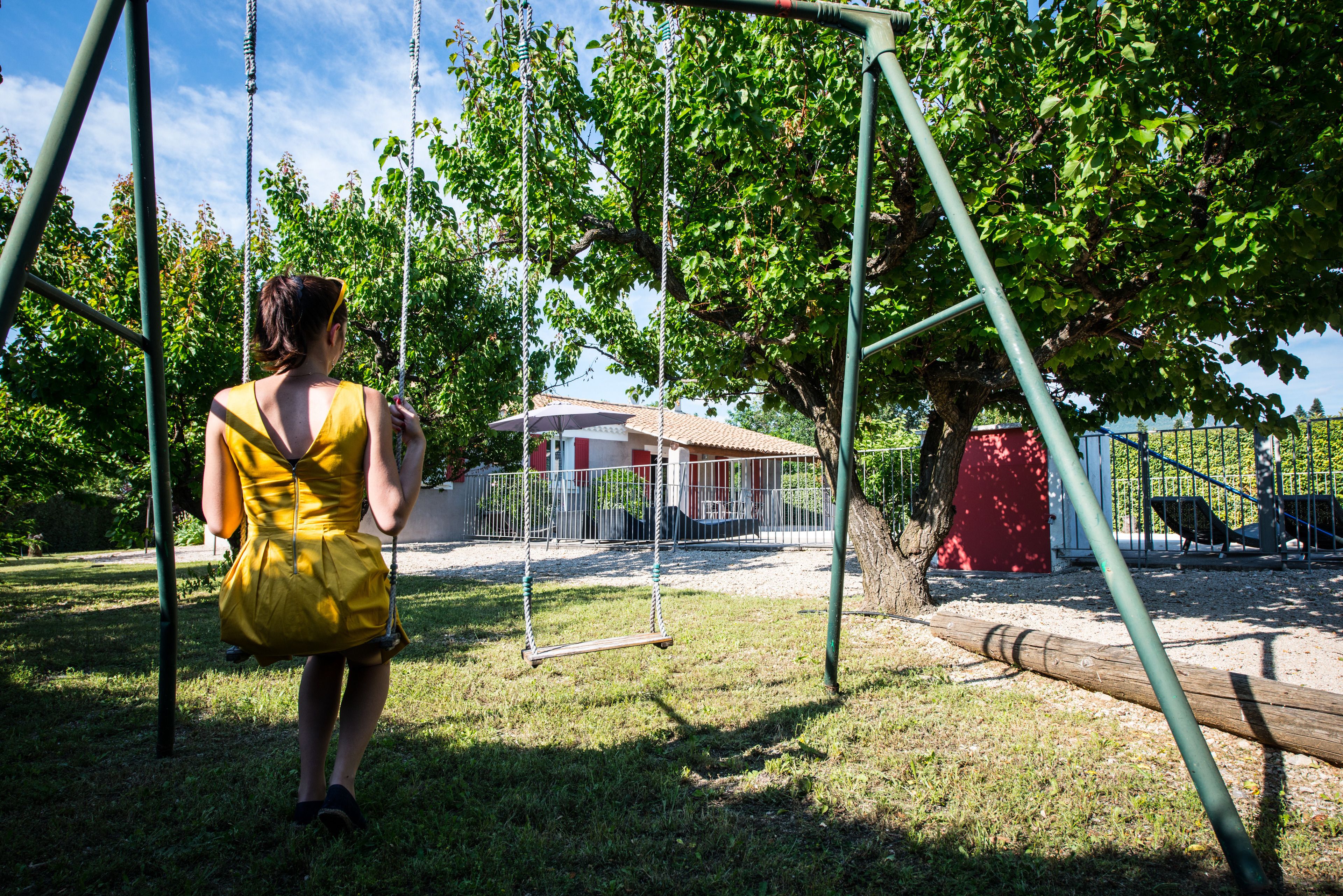 A woman in a yellow dress on a swing set in a park with a red building in the background.