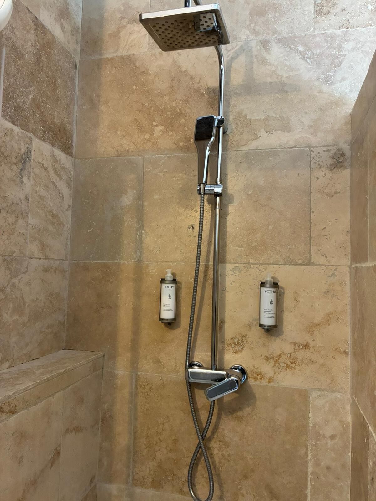 Área de chuveiro com chuveiro manual, controle fixado na parede e dois frascos de sabonete na parede.