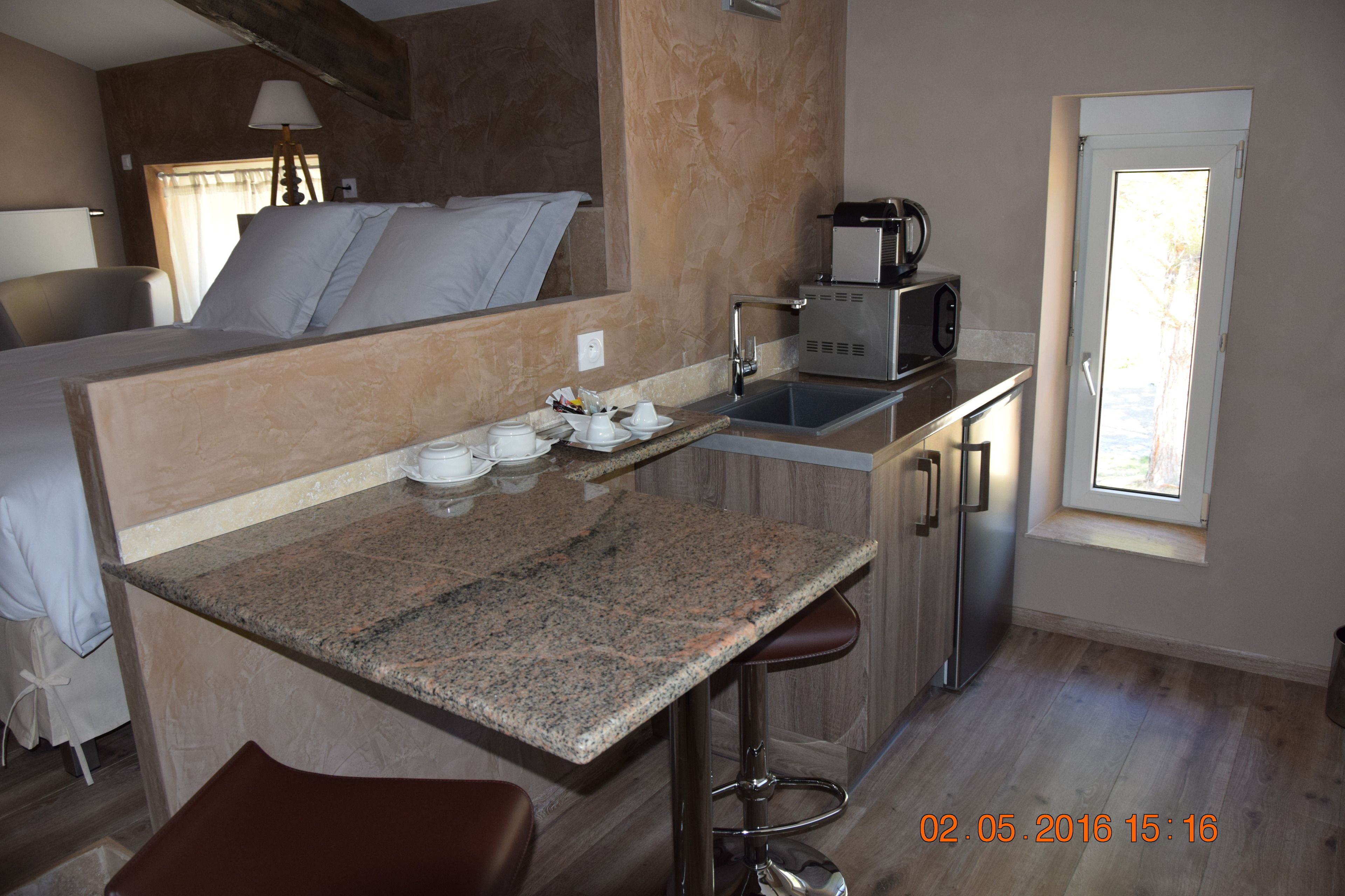 A compact hotel room with a kitchenette, a bed with white linens, a small bar area, and a window providing natural light.