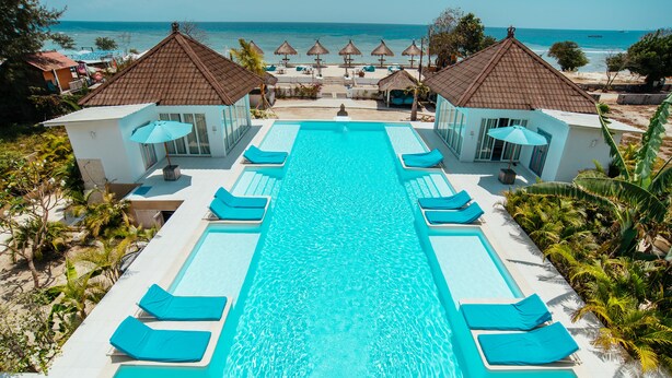 Une piscine rectangulaire à l’eau d’un bleu limpide, entourée de chaises longues et de parasols, avec une vue sur l’océan et la plage en arrière-plan.