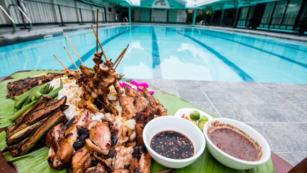 A poolside dining setup with grilled meats, rice, and two dipping sauces on a banana leaf. - Family Country Hotel & Convention Center (General Santos)