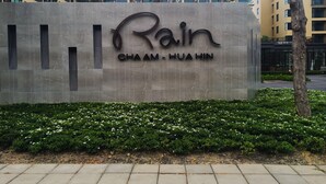 Une enseigne pour Rain Cha Am-Hua Hin, dotée d’un design moderne et agrémentée d’un espace paysager avec des arbustes et des arbres.
