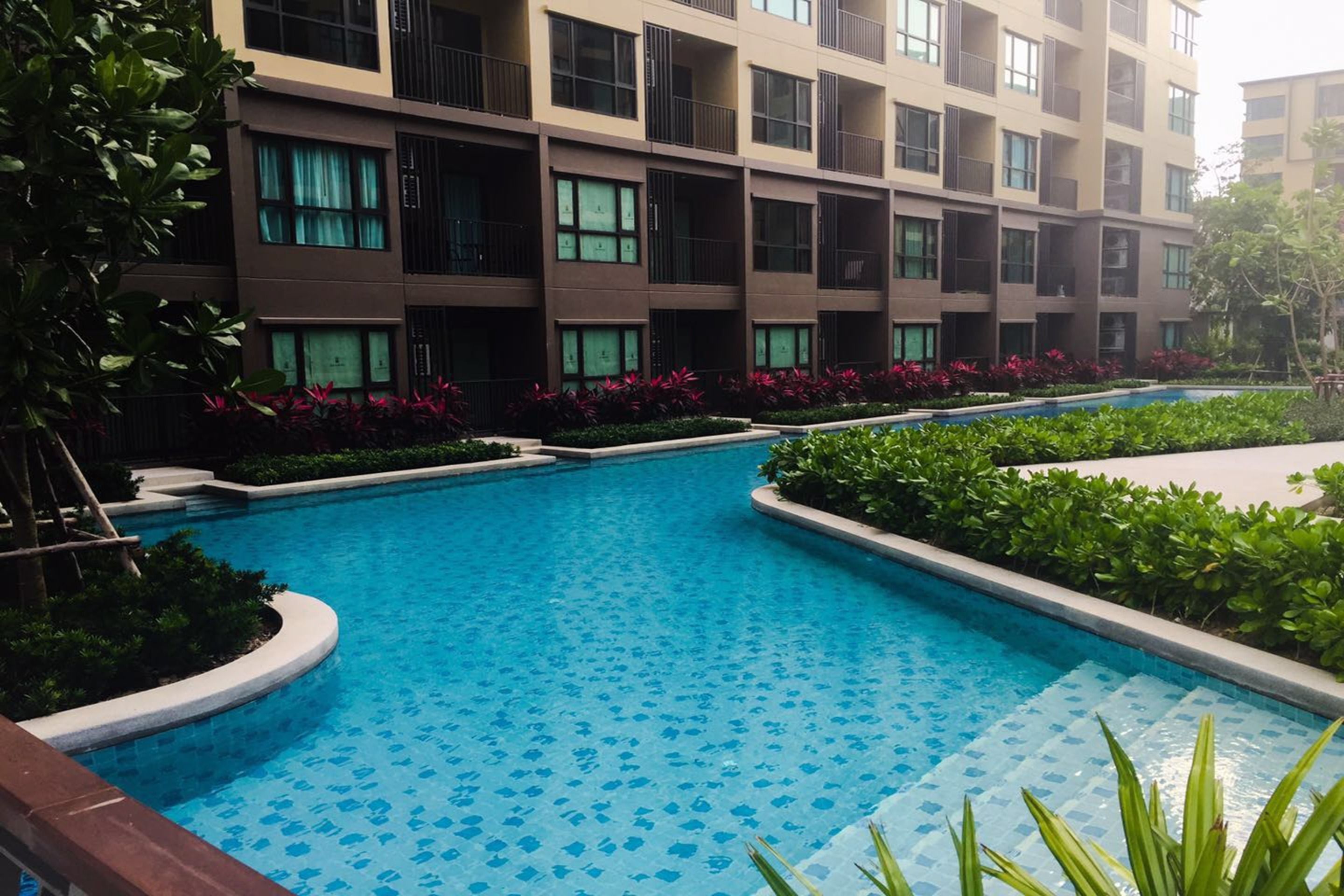 A modern hotel with a swimming pool surrounded by lush greenery and a multi-story building with balconies.