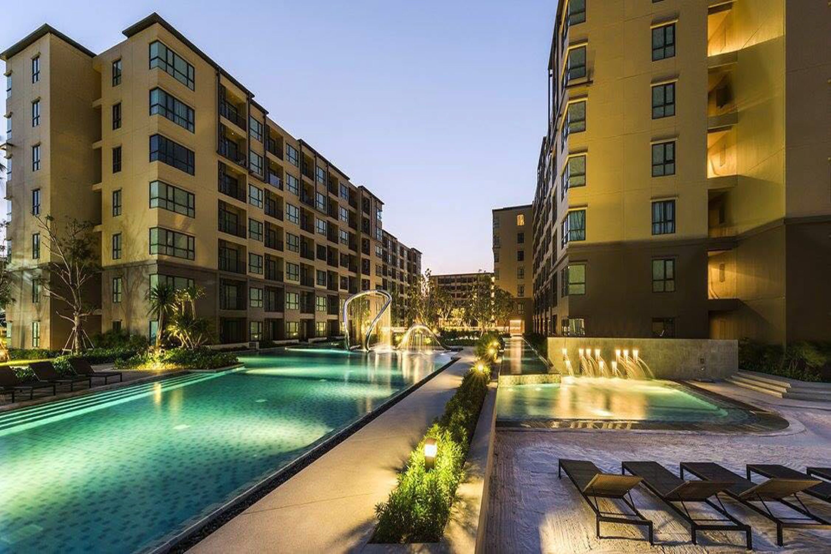 Un hotel con varios pisos, piscina con fuente de agua y tumbonas.