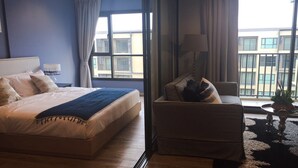Une chambre à coucher avec un lit, une chaise, une lampe et une vue sur les immeubles à travers de grandes fenêtres.