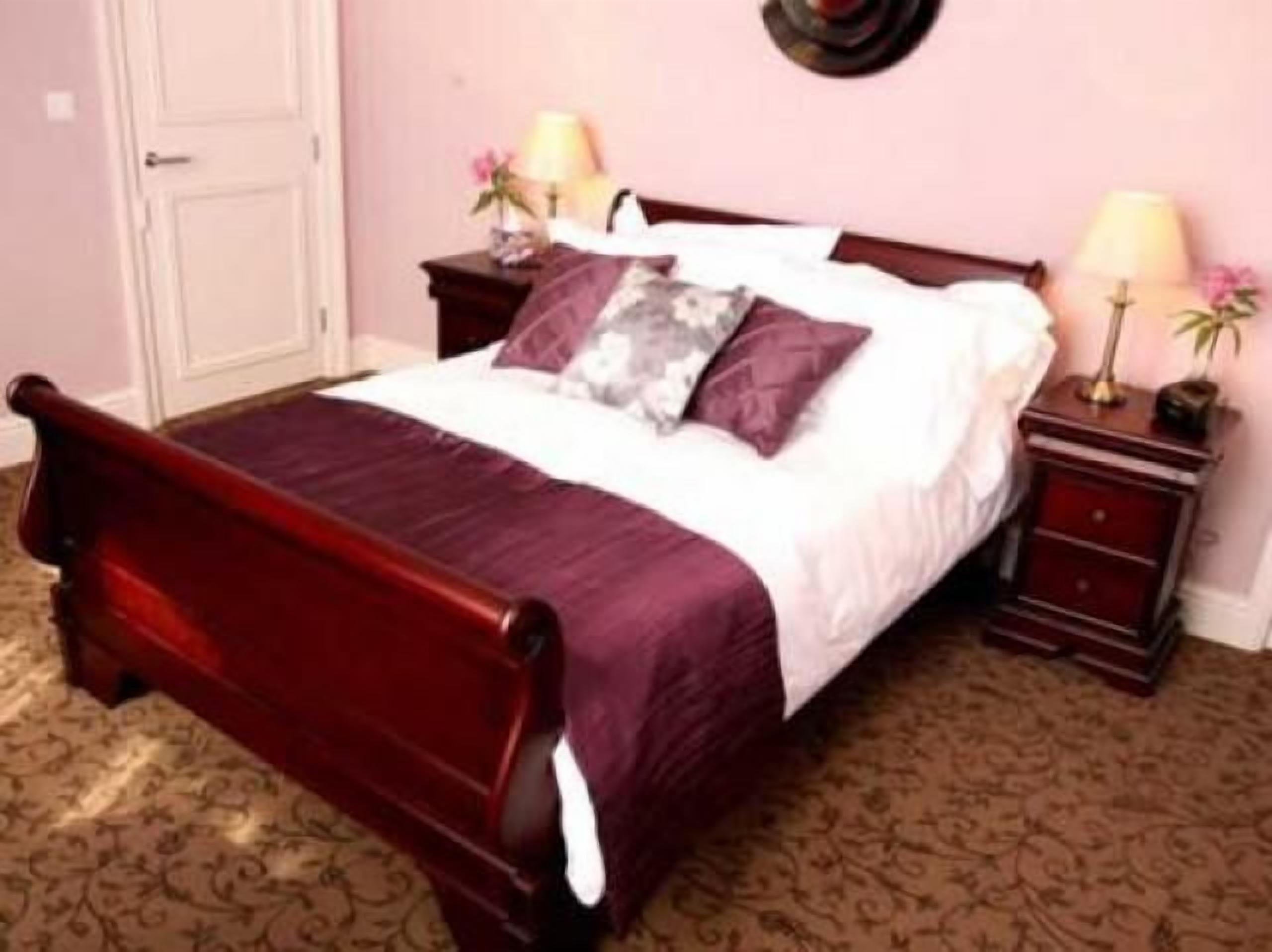 A bedroom with a wooden bed, a red bedspread, two nightstands with lamps, and a wall clock.