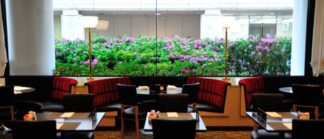 A restaurant with round tables and black chairs, a large window with a view of a garden with pink flowers, and a red and black patterned sofa.