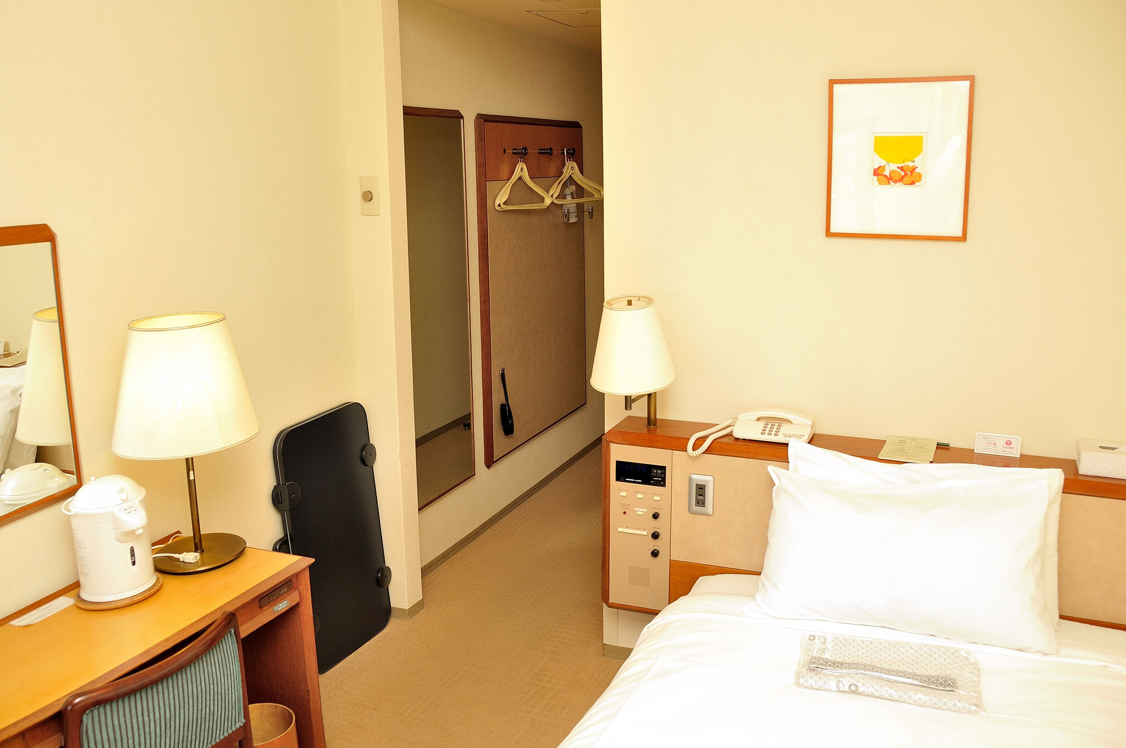 A hotel room with a bed, two bedside lamps, a desk with a lamp, a coffee maker, a telephone, and a coat rack with hangers.