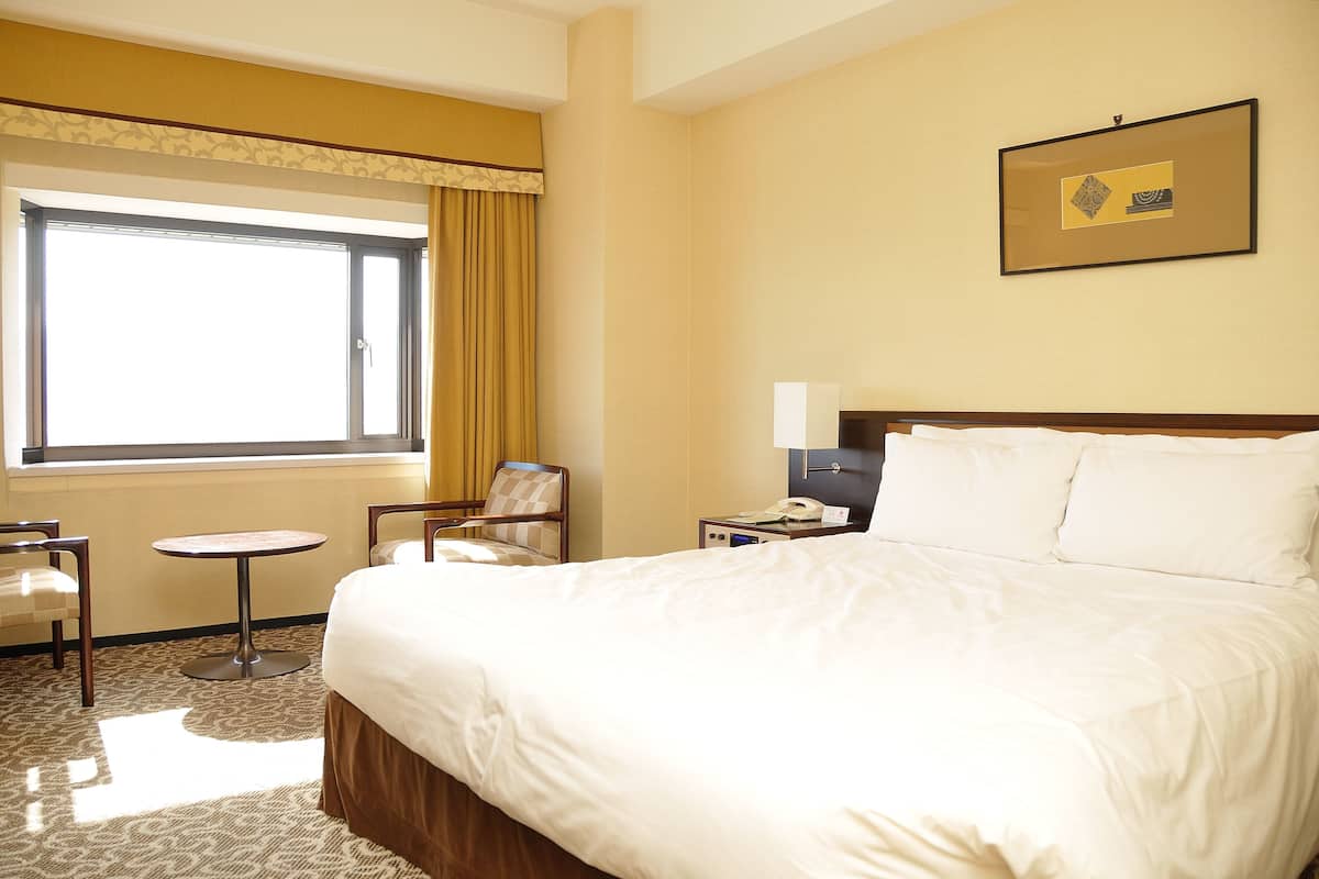 Ein Hotelzimmer mit einem großen Bett, einem Nachttisch mit Lampe, einem Sessel, einem kleinen runden Tisch und einem Fenster mit Vorhängen.
