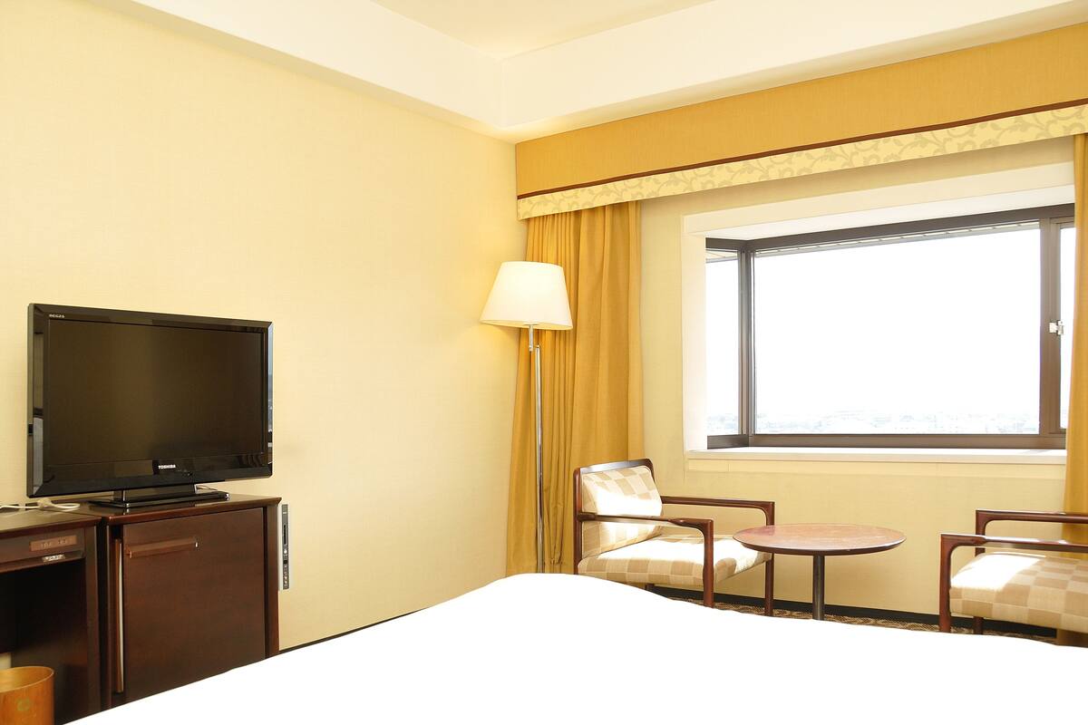 Ein Hotelzimmer mit einem Bett, einem Schreibtisch mit Fernseher, einem Sessel, einem kleinen Tisch und einem Fenster mit Vorhängen.