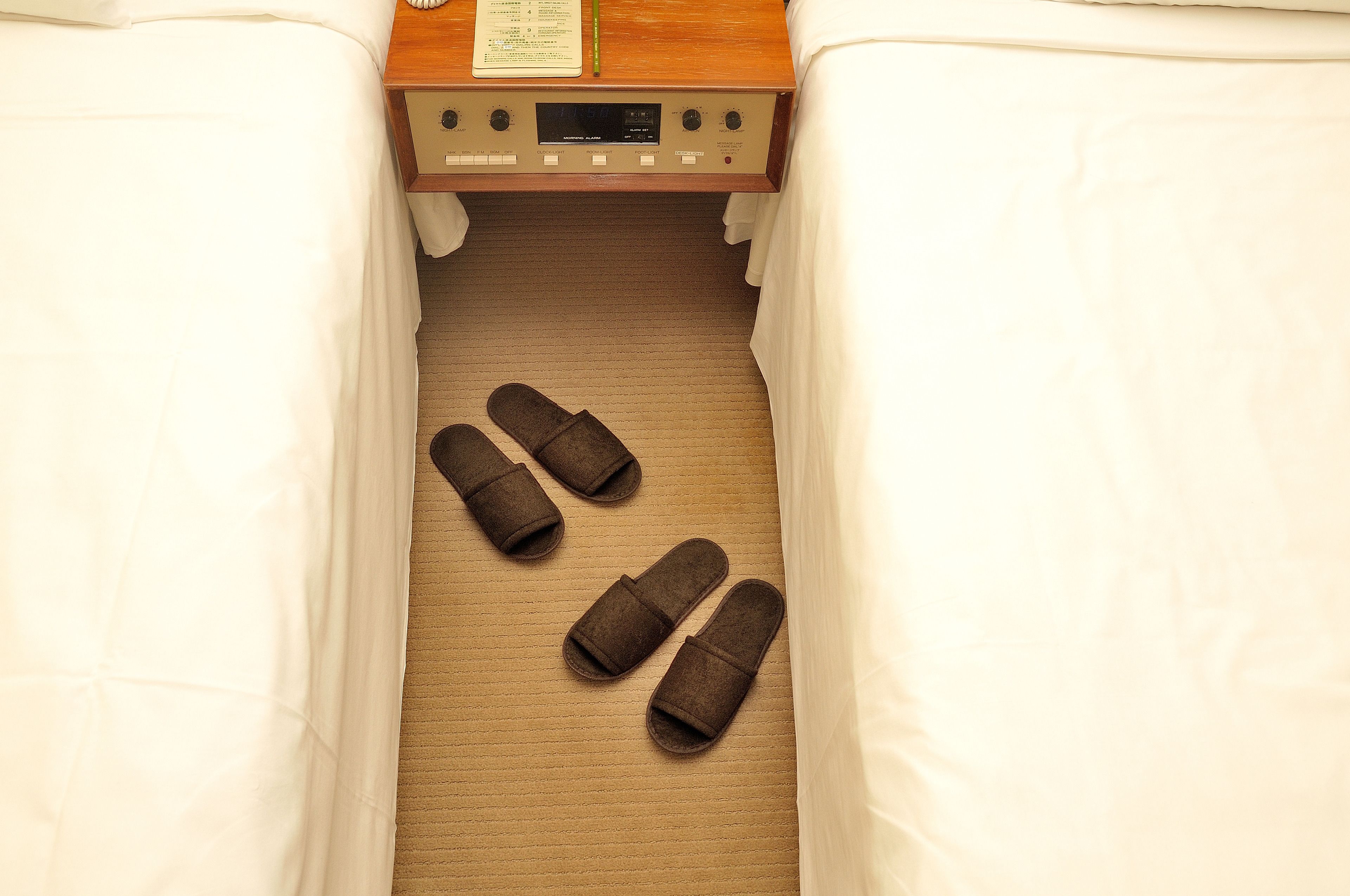 A hotel room with a bed, a wooden nightstand with a clock, and a pair of slippers on the floor.
