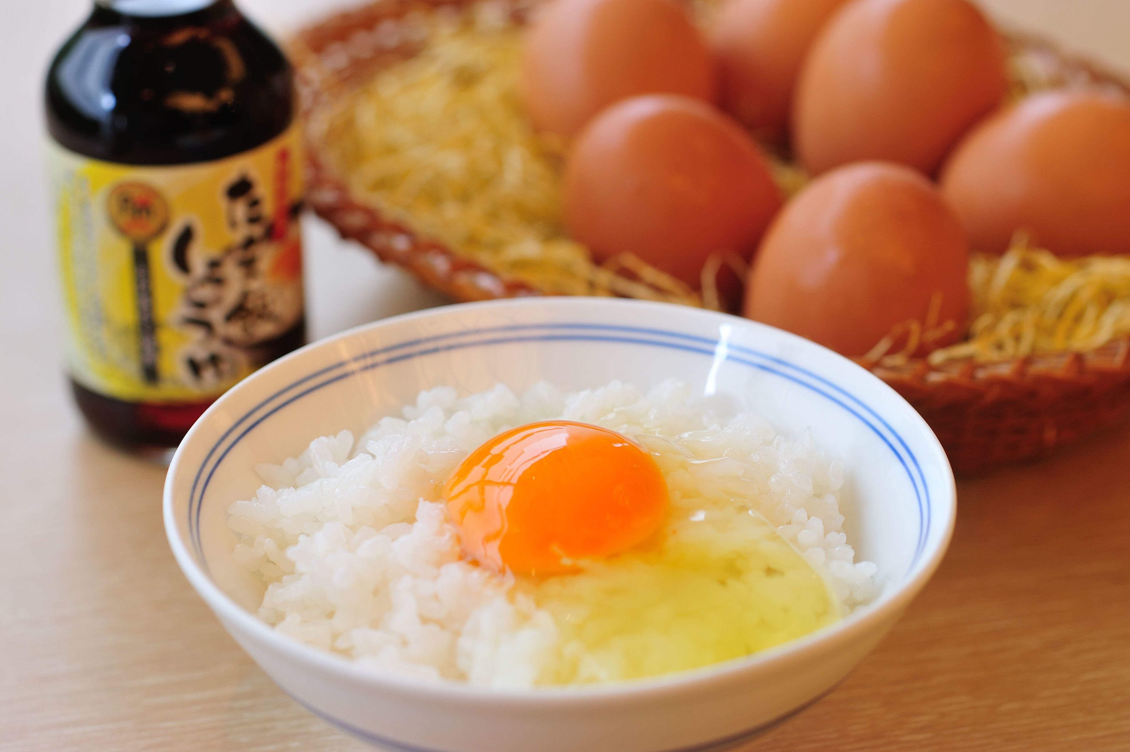 A bowl of white rice with an egg yolk in the center, a bottle with a label, and a basket with eggs in the background.
