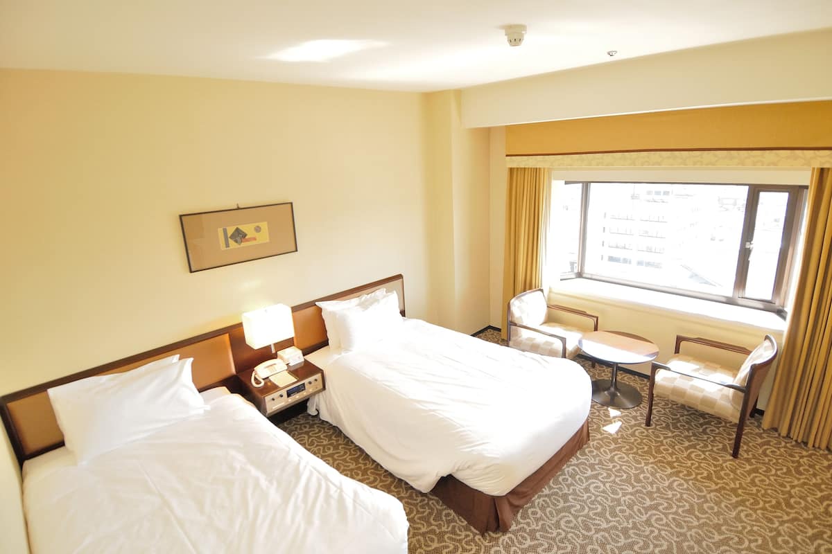 Ein Hotelzimmer mit zwei Betten, einem kleinen Tisch, einem Stuhl und einem Fenster mit Vorhängen.